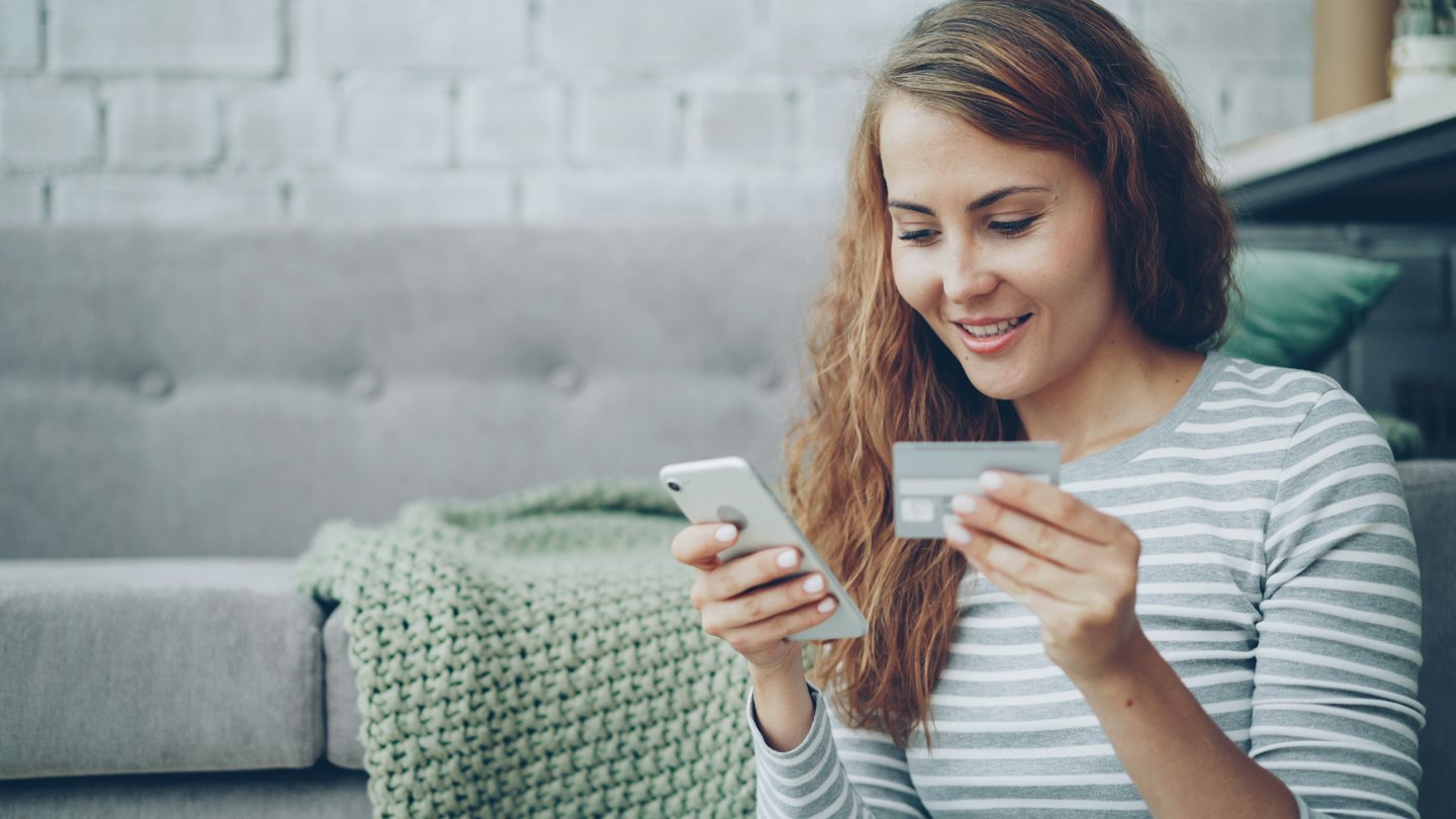 Kobieta uśmiecha się, trzymając telefon i kartę kredytową podczas zakupów online.